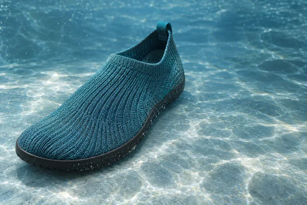 Aqua knitted slip-on water shoe resting on a pale blue-gray submerged floor, with its rounded toe pointing toward the lower left and small bubbles along the…