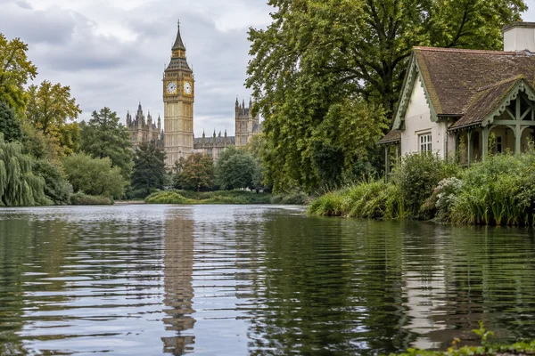 A large pale cottage with a timber porch stands on the right bank of a park lake, while Elizabeth Tower and Gothic buildings rise beyond the trees across the…
