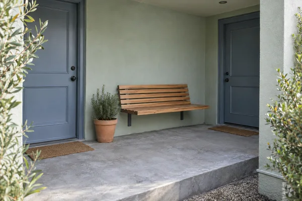 A recessed residential porch with two blue-grey doors, a wall-mounted wooden slat bench, a terracotta pot of rosemary and a broad grey concrete landing.