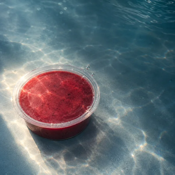 A shallow clear plastic cup of deep red puree rests on a blue-grey underwater surface, lit by a warm patch of light amid rippling caustics; small bubbles sit…