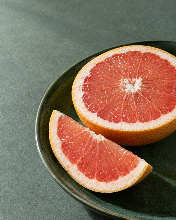 A cut pink grapefruit half and a large wedge rest on a dark green glazed plate, cropped along the right and lower edges.