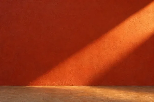 A textured red-orange plaster wall meets a narrow clay-coloured floor, with a broad diagonal band of warm golden light crossing the wall and spilling across…