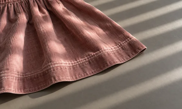A dusty-rose corduroy skirt lies diagonally across a warm-grey tabletop, with its stitched hem and darker band of fabric visible beneath broad parallel shadows.
