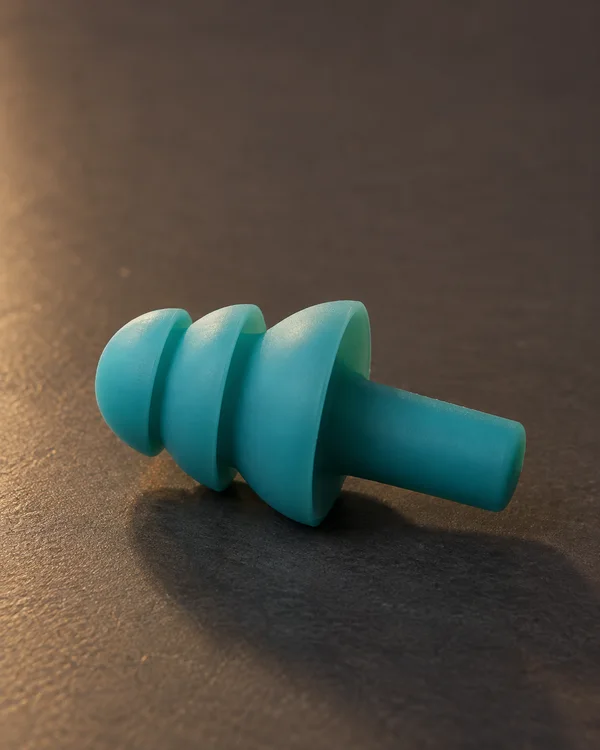 A teal silicone earplug lies sideways on a dark charcoal shelf, with three rounded flanges and its grip stem pointing to the right.