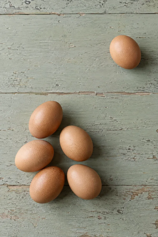 Six brown eggs lie on a worn pale-sage wooden tabletop: five gather in the lower-left area, while one sits apart in the upper right.