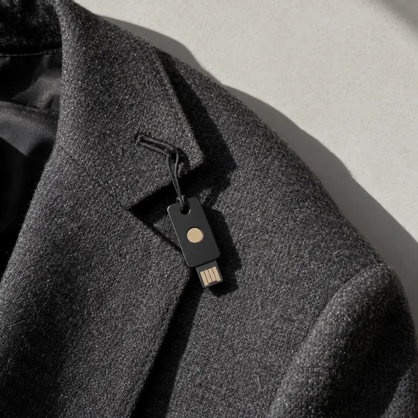 A black rectangular hardware key with a circular gold contact and exposed gold connector hangs by a short black cord from the buttonhole of a charcoal wool…