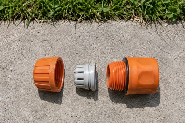 An orange plastic garden-hose connector is arranged in three separated parts on a concrete slab: a threaded collar, pale split sleeve and orange socket body…