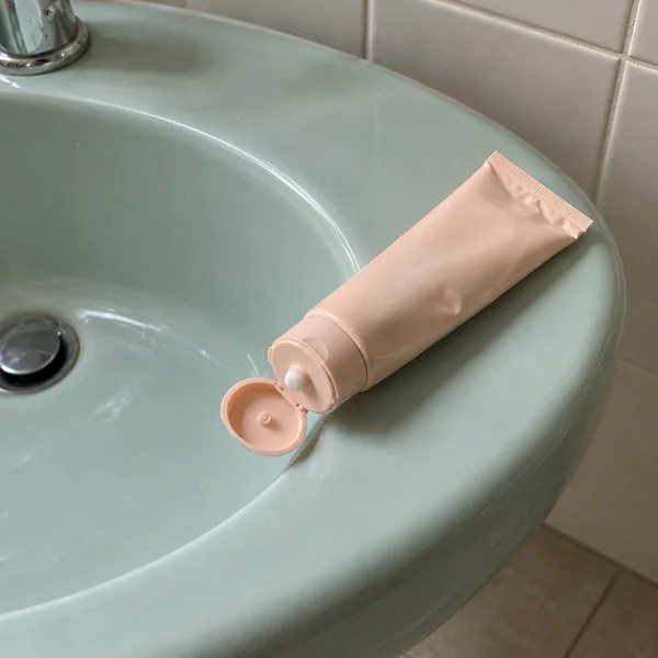 A pale-peach squeeze tube with its flip cap open lies diagonally across the rim of a seafoam ceramic bathroom basin.
