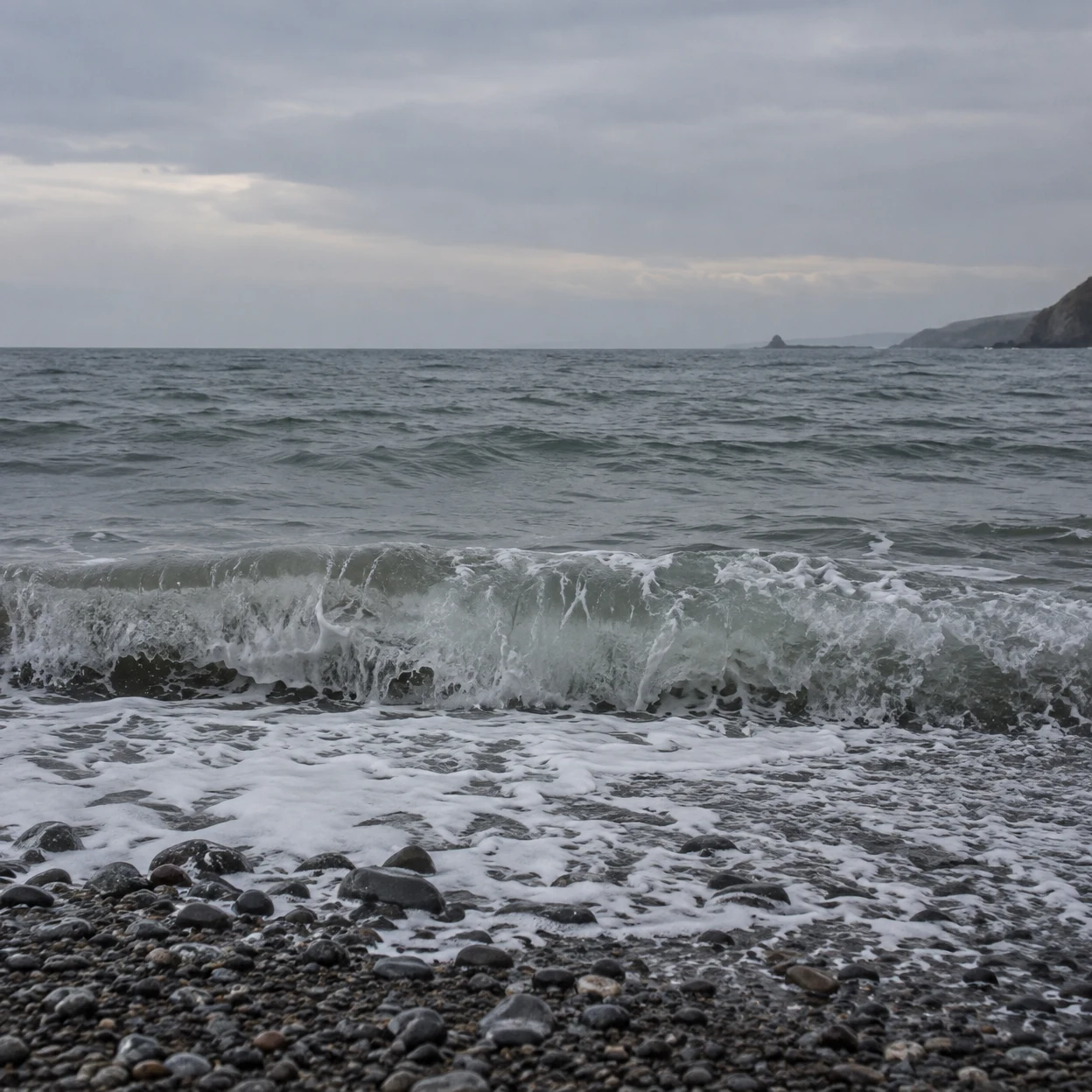 A grey-green wave breaks over rounded dark shingle on a wet beach beneath an overcast slate sky, with a distant headland along the right horizon.