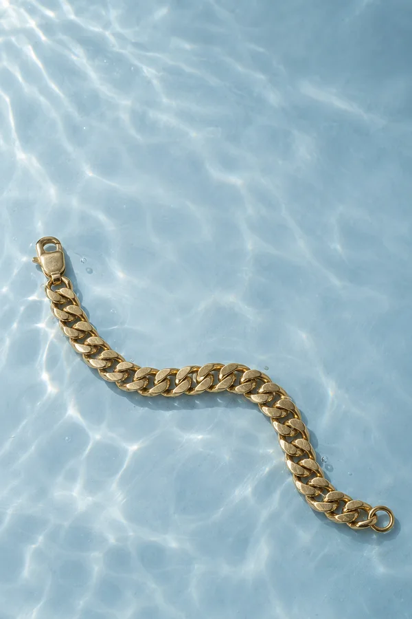 A gold curb-chain bracelet lies in a loose S-curve on a pale blue, rippled underwater surface, with its clasp at one end and a ring at the other; open space…