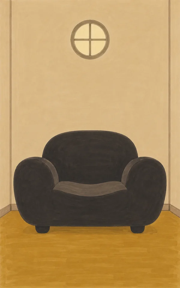 A very wide charcoal armchair with rounded arms and short feet sits centered in a warm ochre-floored room beneath a small circular window.