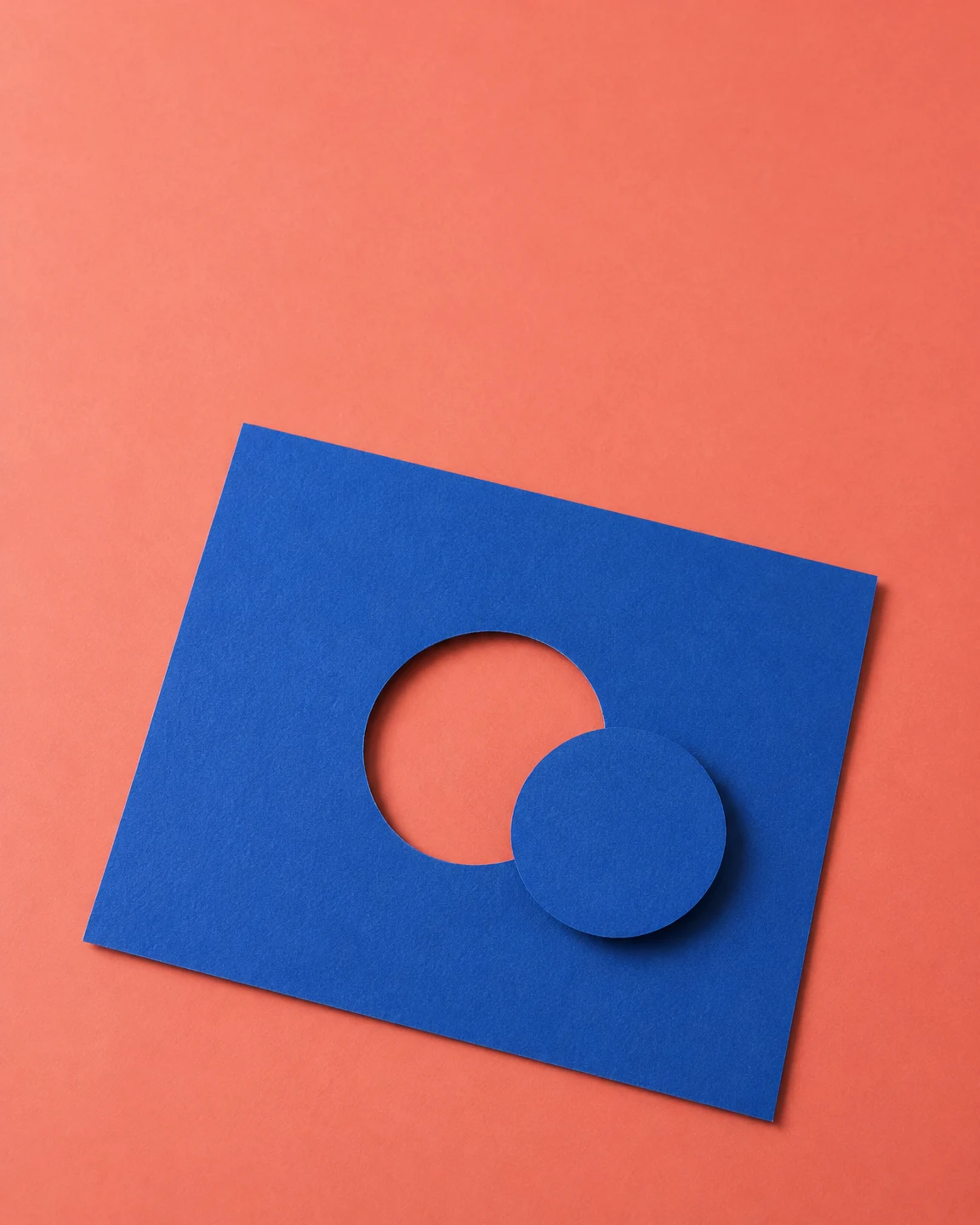 A cobalt-blue rectangular sheet lies at an angle on a coral surface, with a circular cutout and a matching blue disk overlapping the lower-right edge of the…