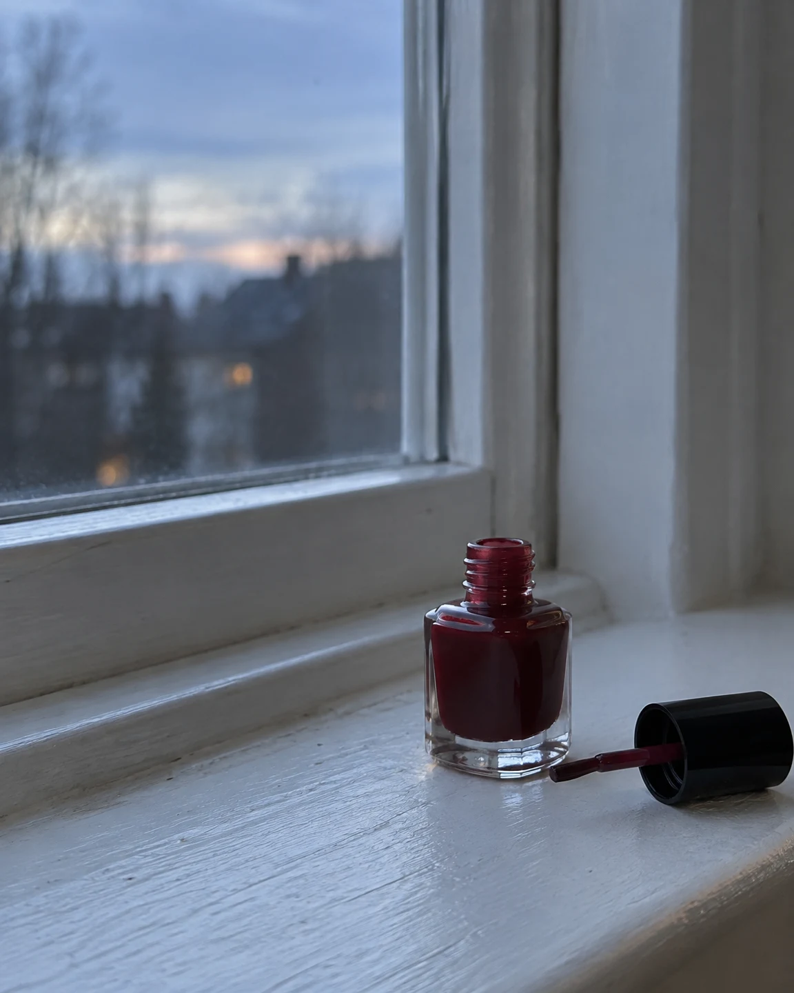 An open, squat clear-glass bottle of deep red nail polish and its black brush cap sit on a pale painted windowsill beside a window, with a softly blurred…