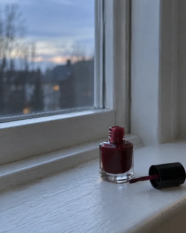 An open, squat clear-glass bottle of deep red nail polish and its black brush cap sit on a pale painted windowsill beside a window, with a softly blurred…