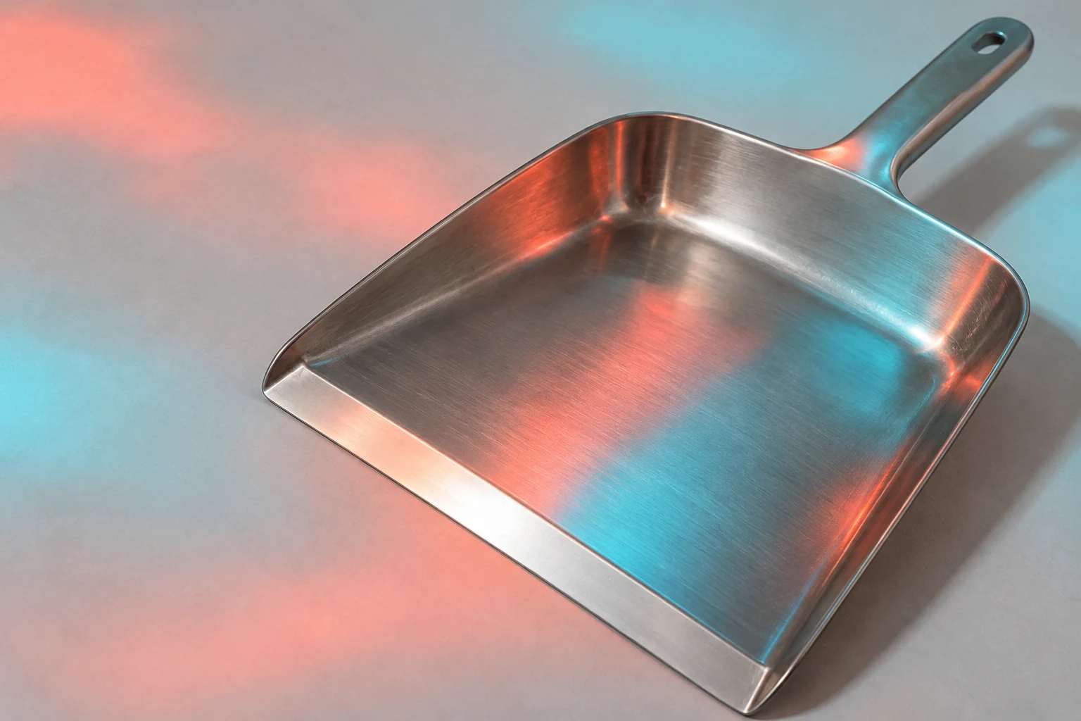 A stainless-steel dustpan lies diagonally on pale grey paper, its broad shallow pan filling most of the frame and its metal handle pointing toward the upper…