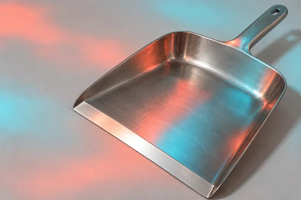 A stainless-steel dustpan lies diagonally on pale grey paper, its broad shallow pan filling most of the frame and its metal handle pointing toward the upper…