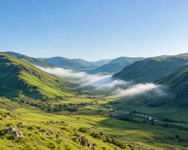 A sunlit grassy valley winds between green and blue-shadowed hills beneath a spacious pale-blue sky, with several soft banks of white mist crossing the valley…