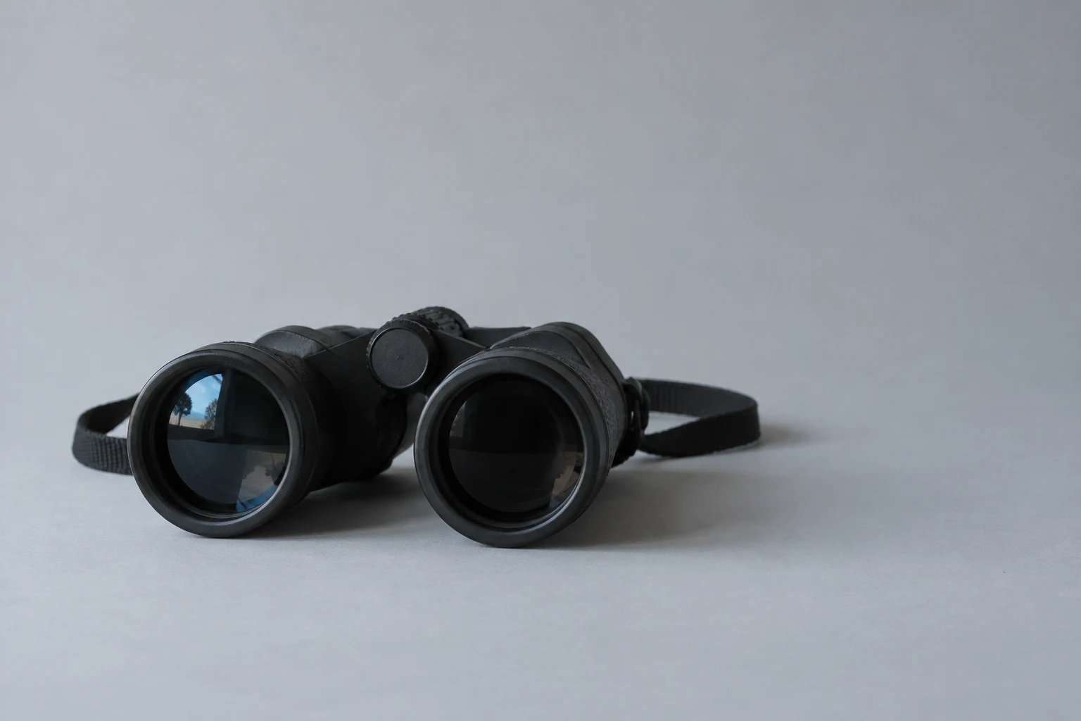 Black rubber-armoured binoculars rest on a pale grey surface, with a woven strap lying behind them.