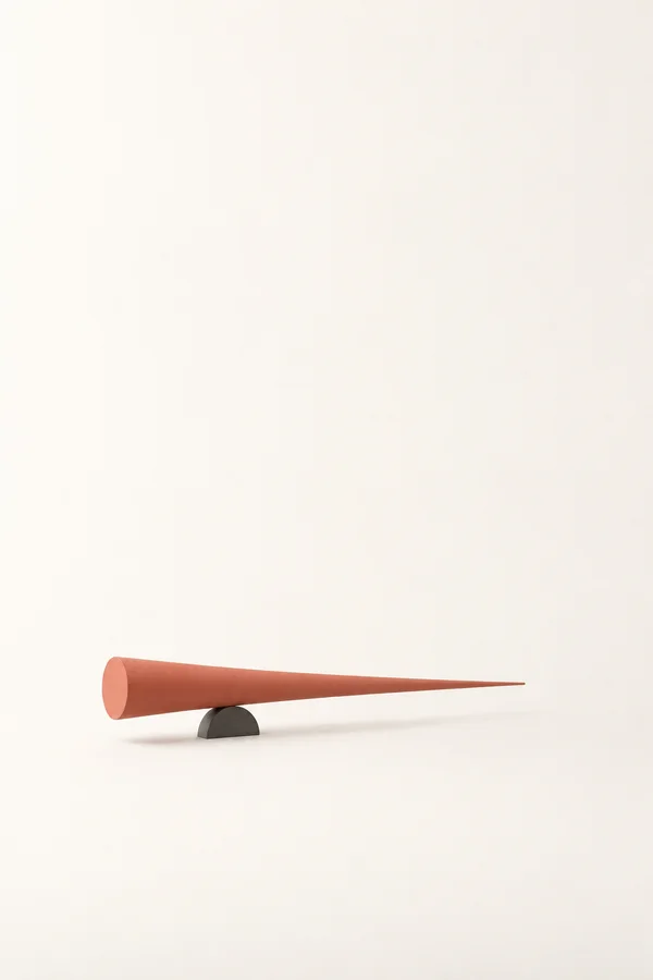 A long muted-coral tapered solid rests horizontally on a small dark graphite half-disc, set low in a warm-white portrait frame with a soft shadow and broad…