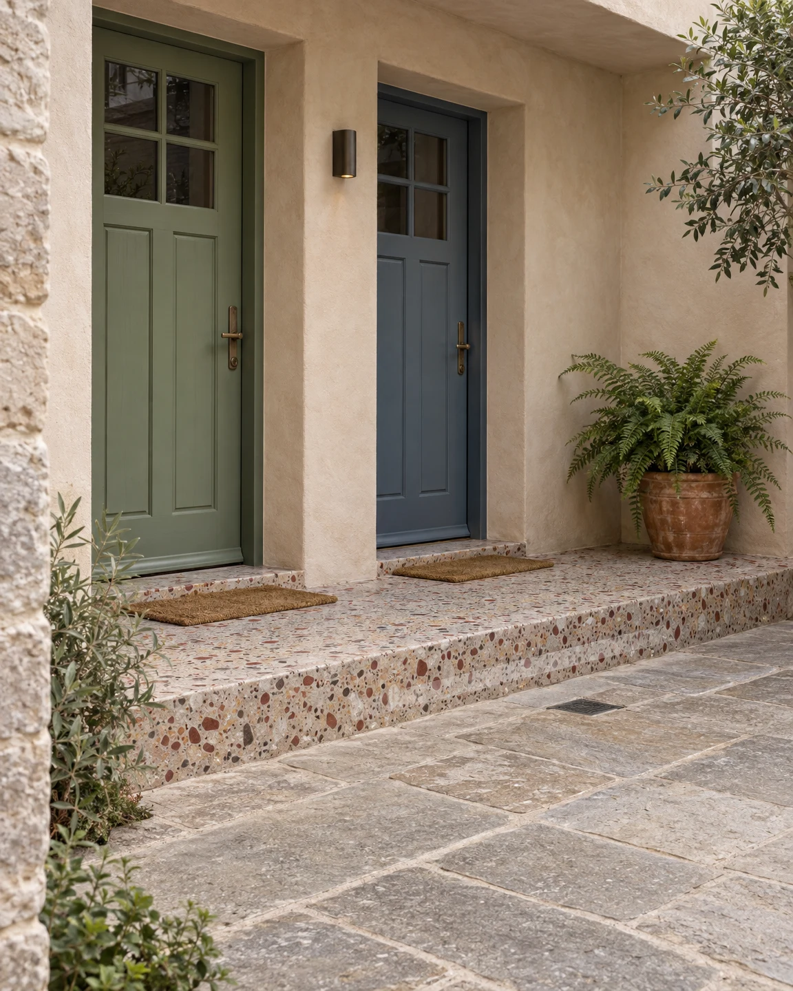 Two neighbouring homes with muted sage and slate-blue paneled doors sit beneath a pale clay-colored covered entrance.