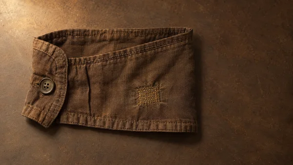 A brown cotton garment cuff lies open on a textured umber tabletop, with a four-hole button at the left and a small rectangular gold-thread repair near the…
