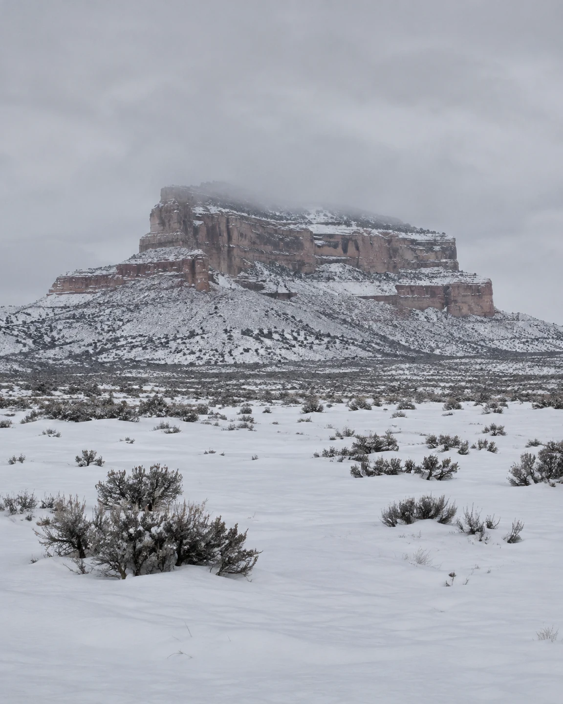 A rust-colored, flat-topped mesa rises above a snow-covered high-desert plain, with snow lining its rock ledges and low mist softening the upper rim.