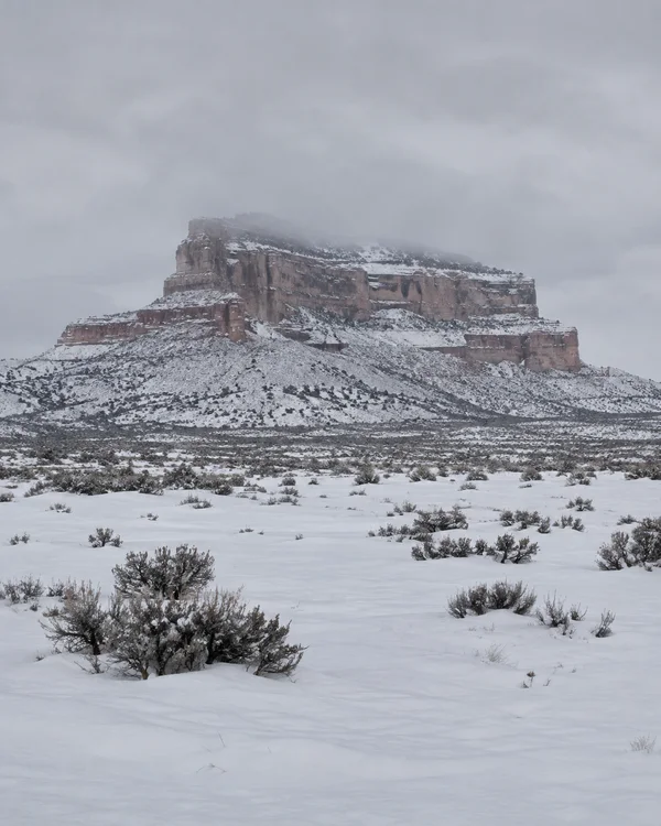 A rust-colored, flat-topped mesa rises above a snow-covered high-desert plain, with snow lining its rock ledges and low mist softening the upper rim.