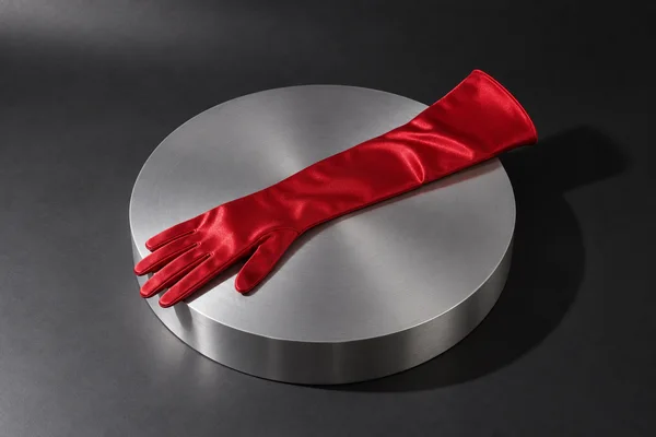 A scarlet satin opera glove lies diagonally across a circular brushed-aluminium turntable against a charcoal studio backdrop, with a strong shadow cast to the…