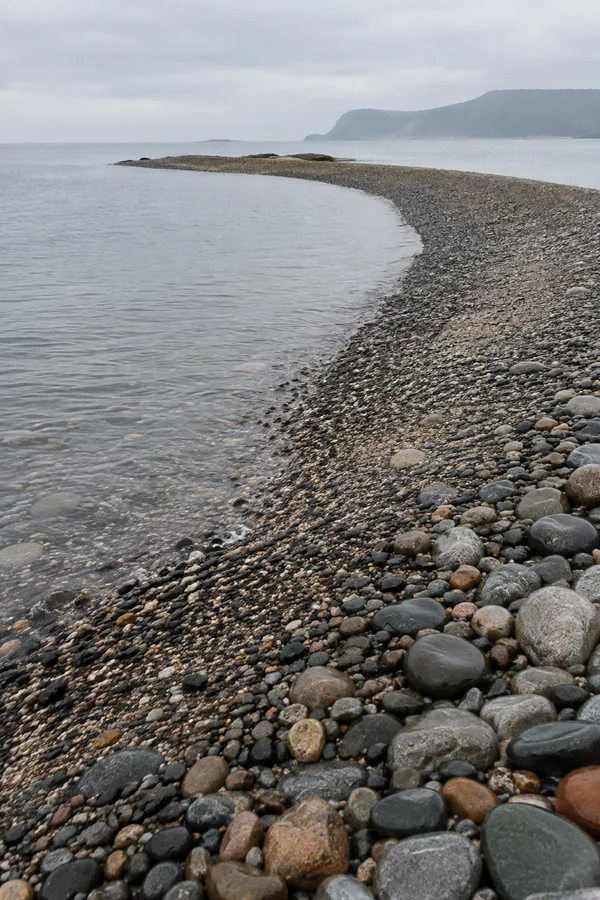 A curved shingle spit of wet pebbles extends beside still grey water under a pale overcast sky, with a hazy headland in the distance.