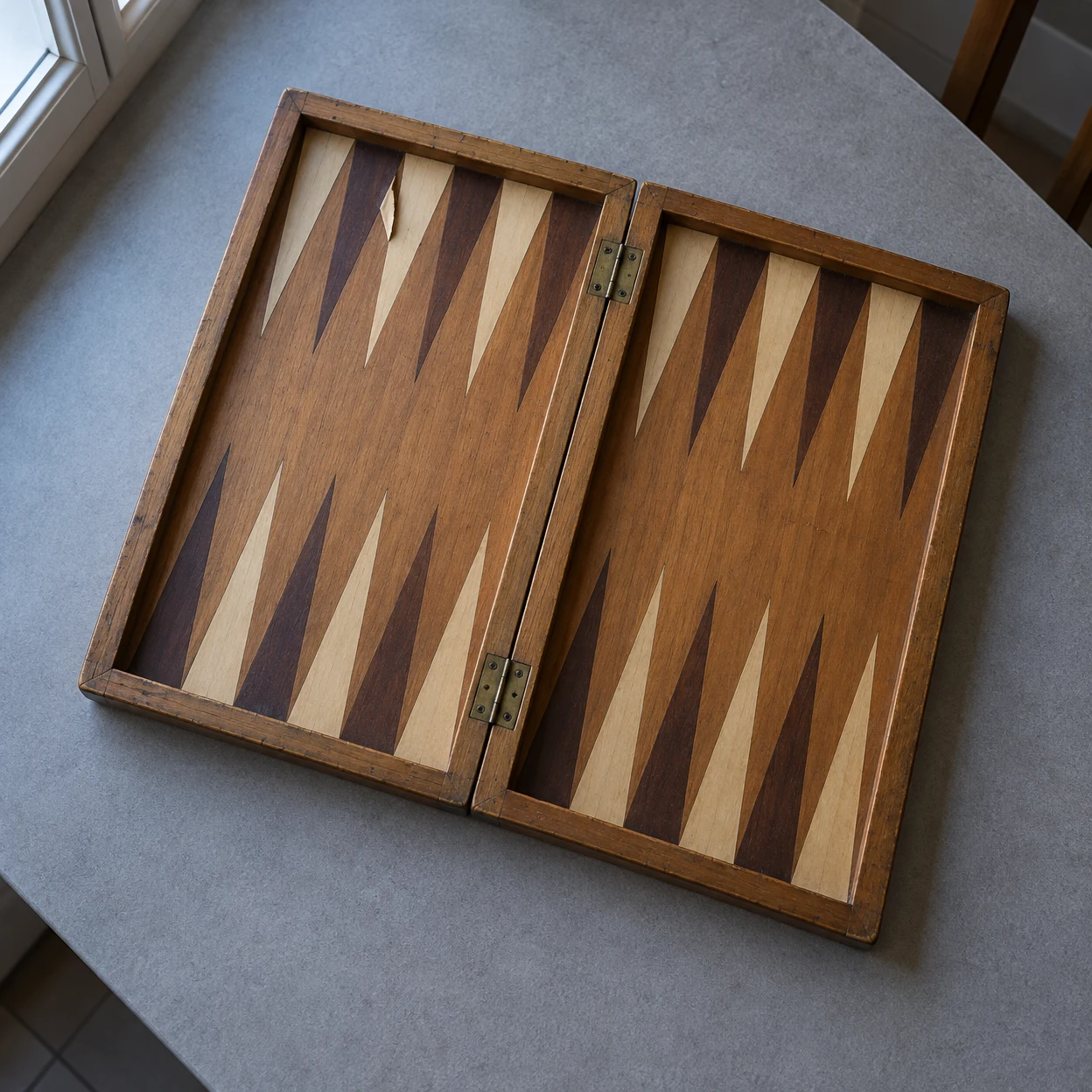 An open, worn wooden backgammon board lies diagonally on a grey tabletop, with alternating light and dark triangular inlays, brass hinges and a small lifted…
