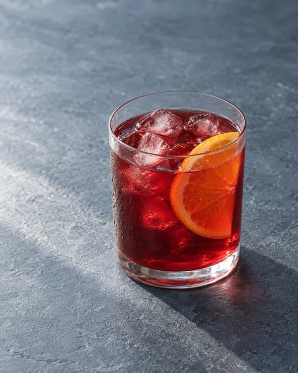 A clear tumbler of ruby-red iced tea with several ice cubes and an orange slice sits on a blue-grey slate tabletop, with open textured space above and to the…
