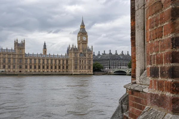 The Elizabeth Tower and Palace of Westminster stand across the Thames beneath a cloudy sky, with a weathered red-brick and pale-stone wall filling the right…