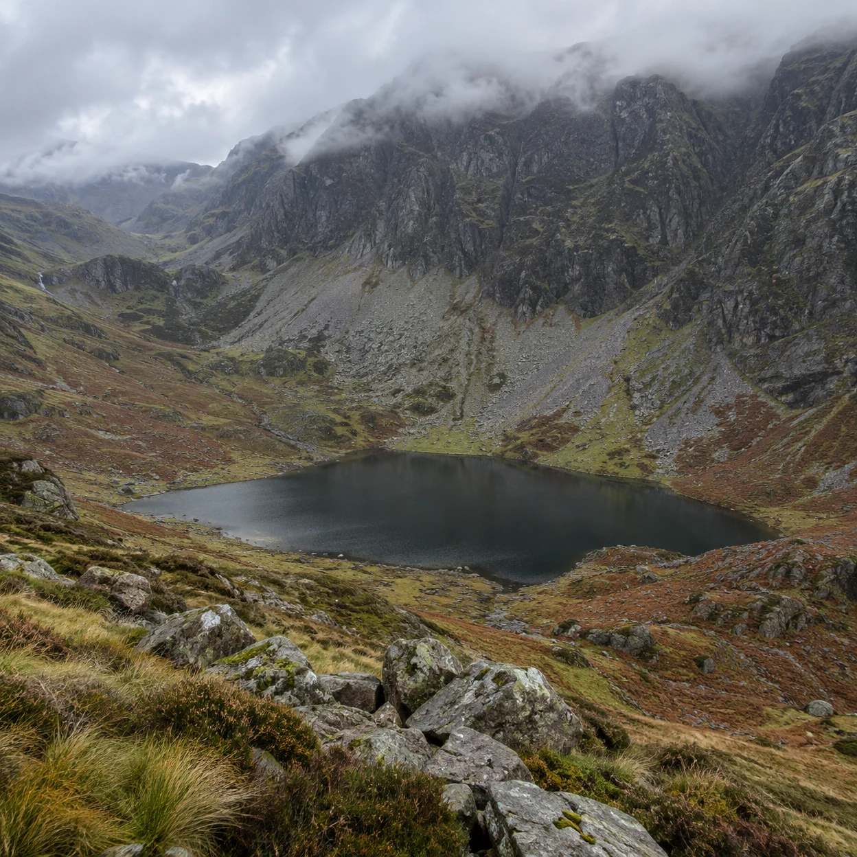 A dark still tarn sits in a steep mountain basin ringed by pale scree slopes and russet moorland, with wet boulders and tussock grass in the foreground and low…