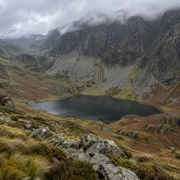 A dark still tarn sits in a steep mountain basin ringed by pale scree slopes and russet moorland, with wet boulders and tussock grass in the foreground and low…