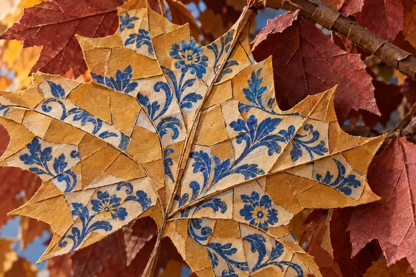 An ochre maple leaf made from overlapping textured paper pieces fills most of the frame, decorated with blue floral scrollwork and surrounded by russet and…