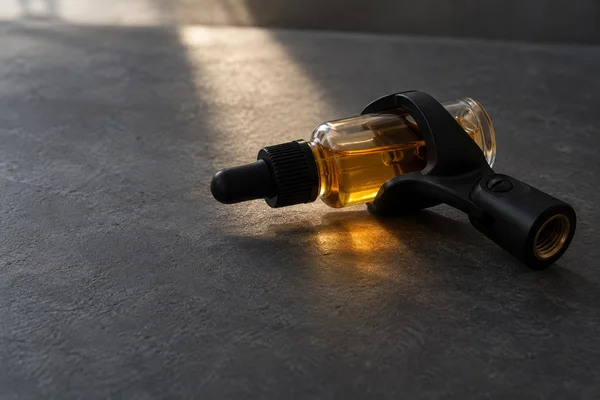 A clear glass dropper bottle partly filled with amber liquid lies in a black microphone clip on a slate-grey tabletop, with a warm beam of light and amber…