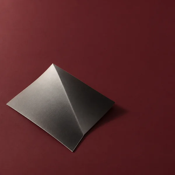 A sharply creased silver-gray sheet forms two angular facets on a muted burgundy field, with a soft shadow falling to the right.