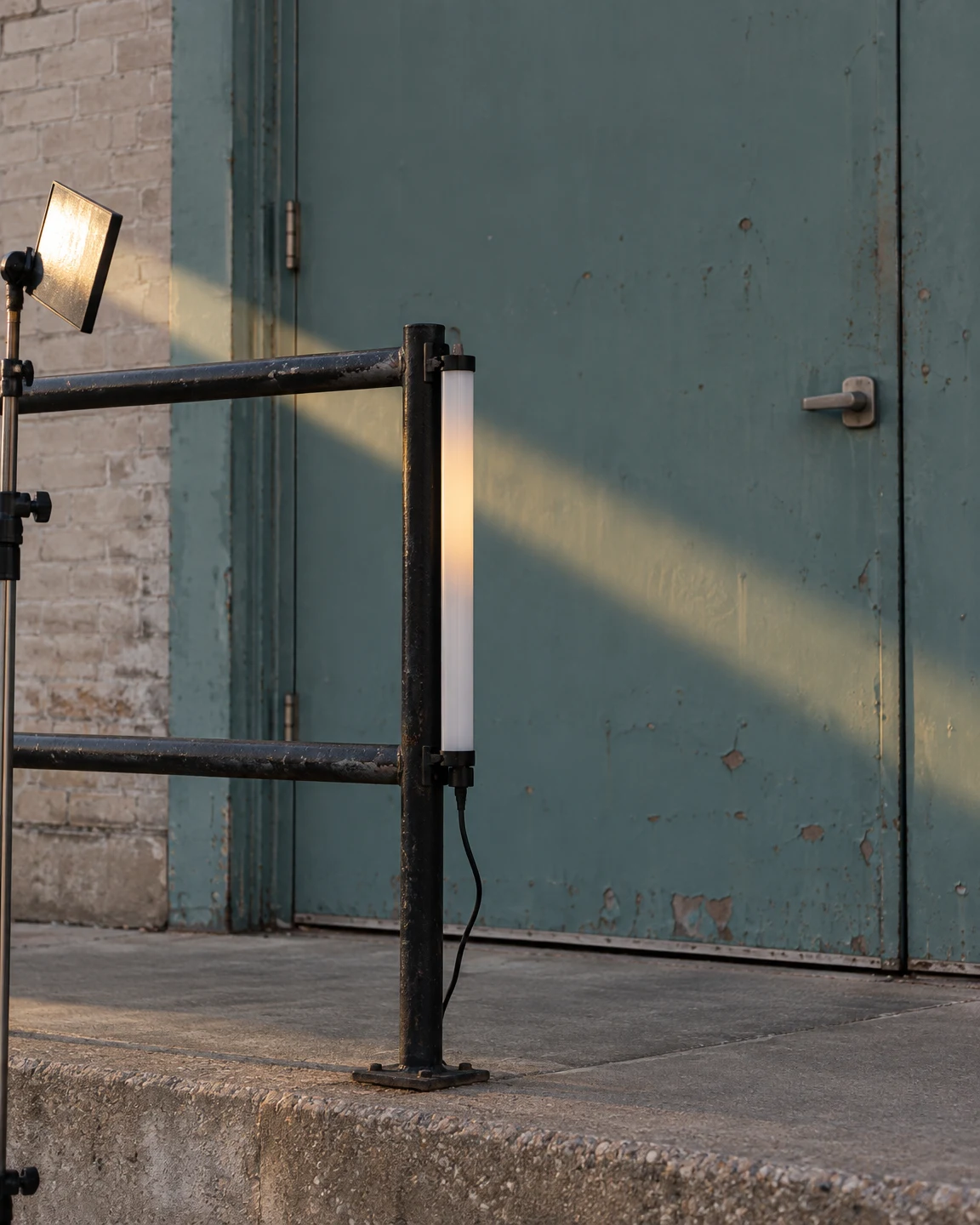 A vertical white LED tube is clipped to a black metal railing in front of a weathered teal loading door.