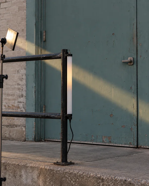 A vertical white LED tube is clipped to a black metal railing in front of a weathered teal loading door.