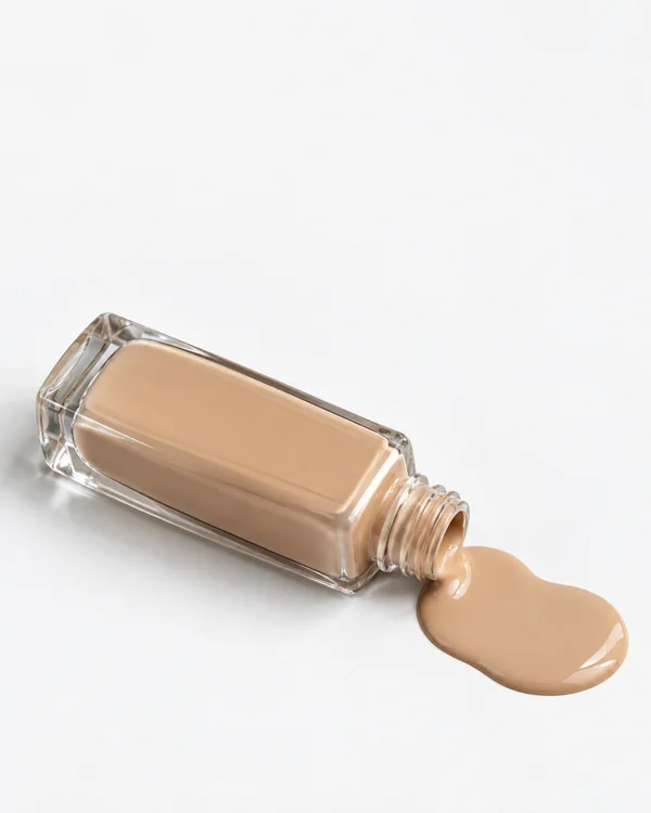 An unlabelled clear glass foundation bottle lies diagonally on a white tabletop, with its open neck beside a glossy pool of warm beige liquid.