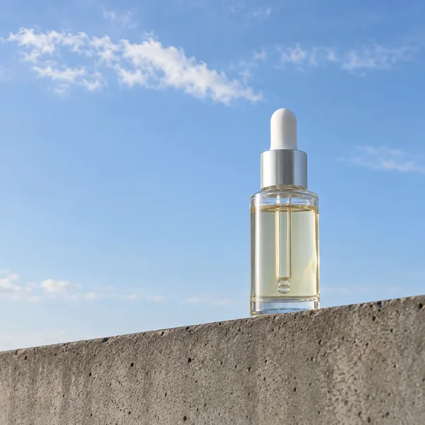 A clear glass dropper bottle filled with pale straw-coloured liquid stands on the sloping top edge of a rough concrete wall against a blue sky with wispy…