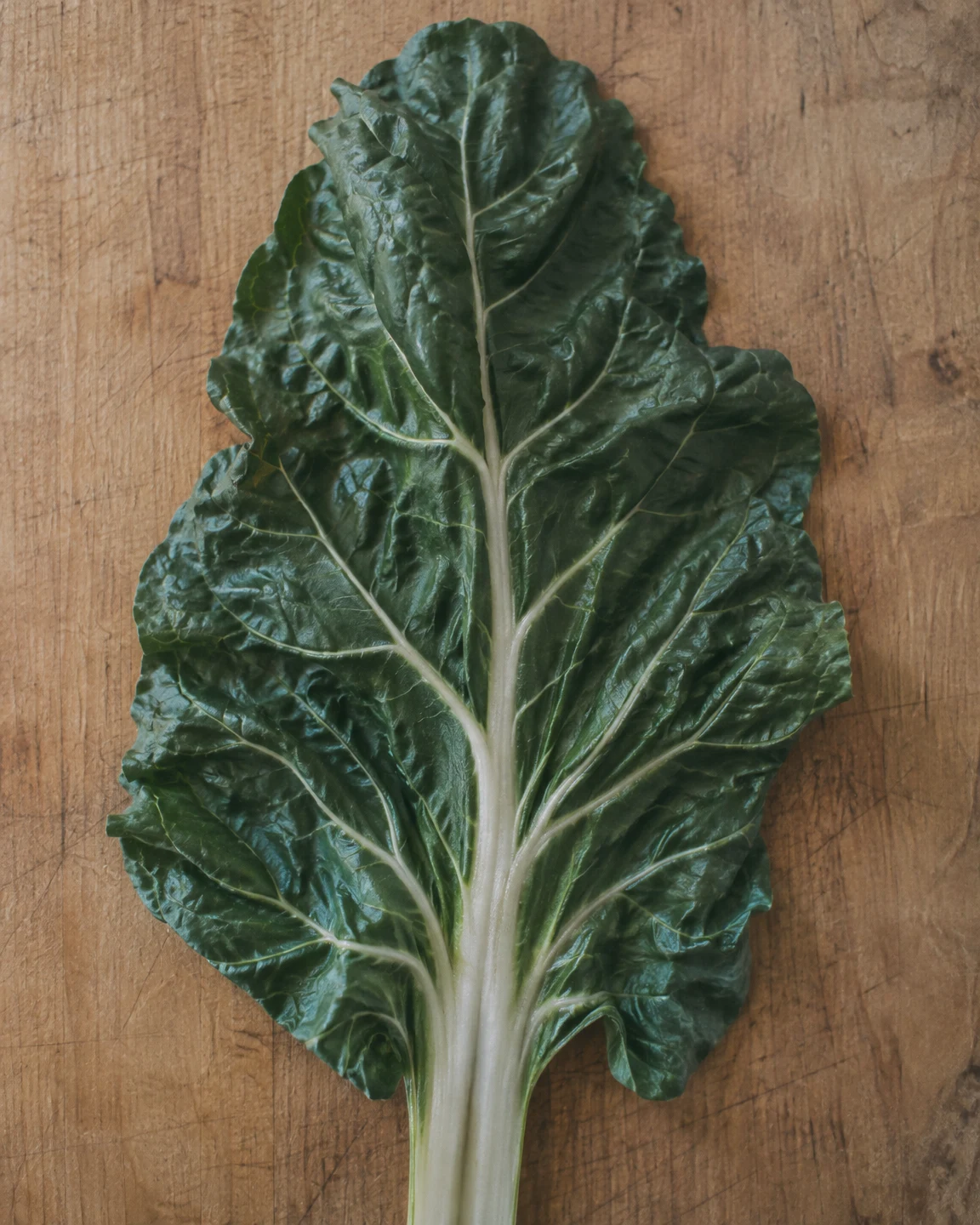 A broad dark-green chard leaf lies on a honey-brown wooden cutting board, its pale central rib extending down past the bottom edge.