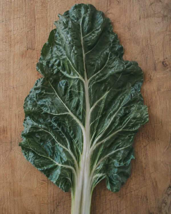 A broad dark-green chard leaf lies on a honey-brown wooden cutting board, its pale central rib extending down past the bottom edge.