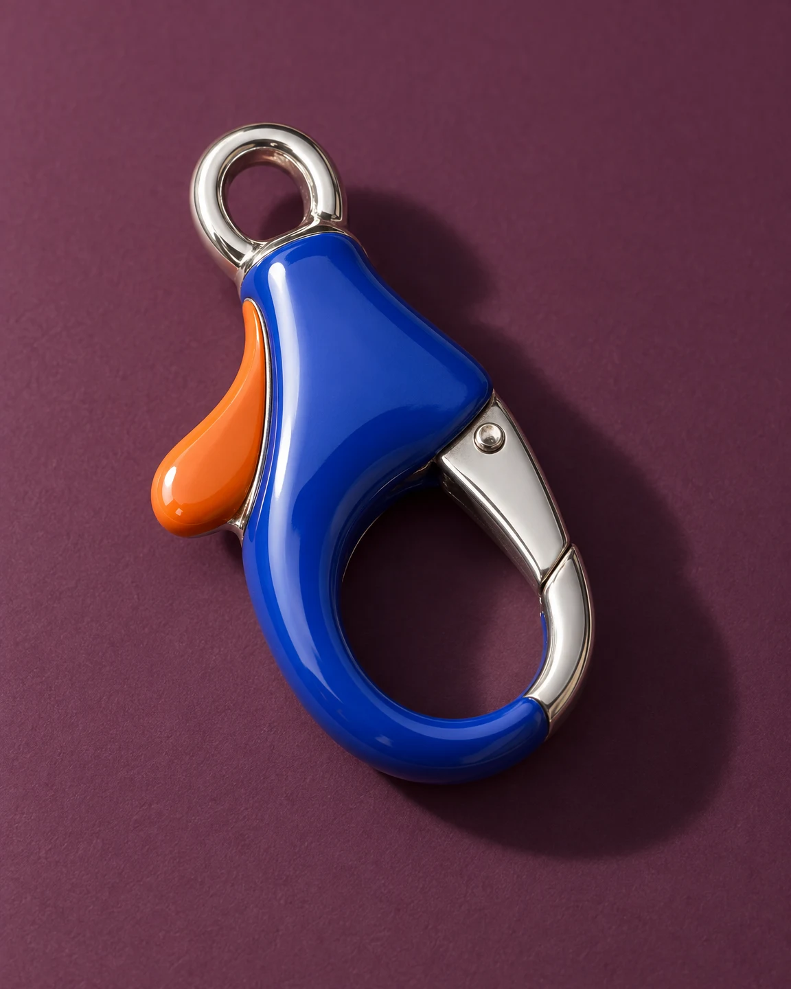 A glossy cobalt-blue and orange jewellery lobster clasp rests diagonally on a plum surface, with a polished silver attachment eye and gate casting a shadow to…