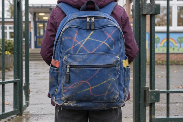 A worn cobalt-blue backpack fills the center of the frame on a child in a plum jacket and charcoal trousers, standing at a metal school gate.