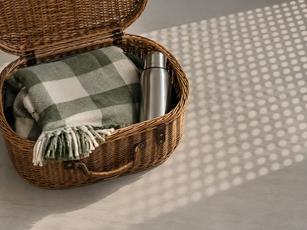 An open oval wicker picnic basket sits on a pale tabletop, holding a folded sage-and-cream checked blanket and a stainless-steel flask.