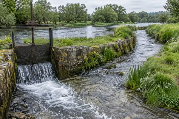 A stone-lined leat curves through a green riverside meadow, with water spilling through a dark iron sluice in the foreground and a broad river and tree-lined…