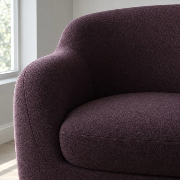 Close-up of an aubergine upholstered armchair beside a bright window, showing the curved back, rounded arm, inset seat cushion, and fabric seams.