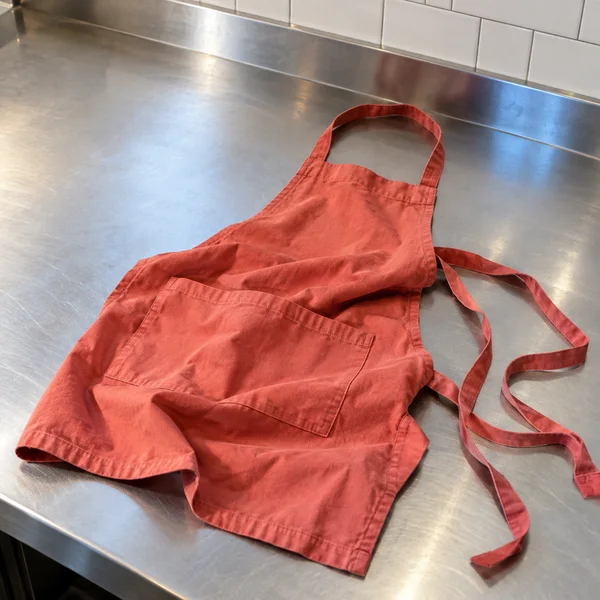 A coral-red cotton apron lies loosely folded on a stainless-steel café prep counter, with its straps trailing across the right side.