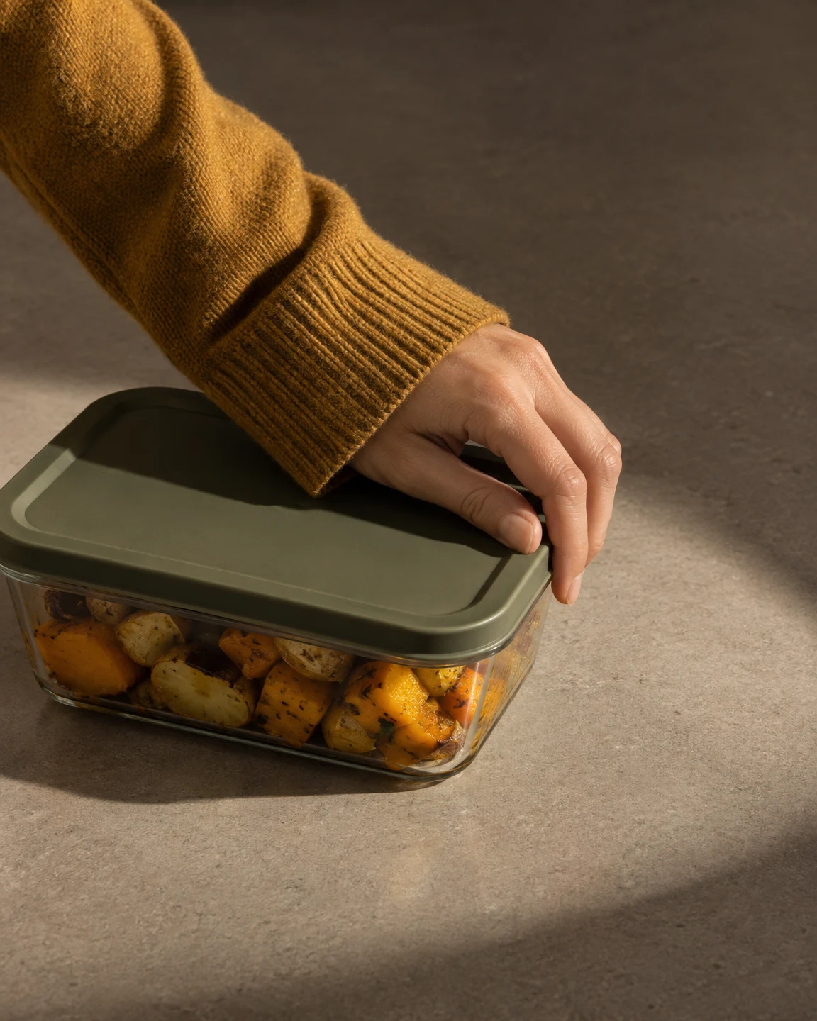A hand in a mustard knit sleeve presses the olive lid of a clear food container filled with roasted squash and potatoes on a warm-grey counter.